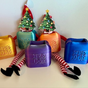 May include: Four square Nee Doh containers in yellow, teal, purple, and blue, each featuring the embossed text Nee Doh. Two elf dolls with red and white striped legs and green Christmas tree hats are positioned behind the containers.