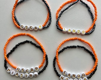 May include: Four beaded bracelets with names spelled out in white letter beads. The bracelets are made of orange and black beads. The names on the bracelets are "OSCAR", "LANDO", "OSCAR PIASTRI", and "LANDO NORRIS".