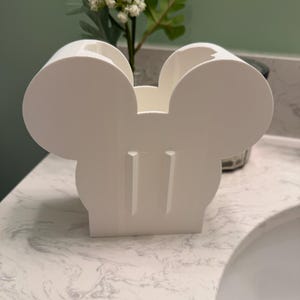 May include: A white plastic organizer featuring a silhouette shape with two large circular ears and two vertical rectangular cutouts on the front face. The container has an open top and a flat base, designed to hold items in a upright position.