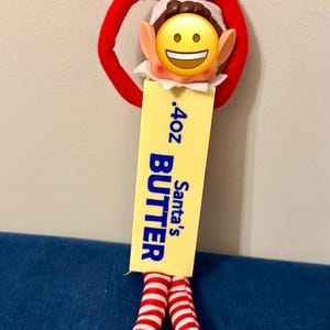 May include: A plush elf doll wearing a red and white hat and red and white striped leggings with black shoes. The doll holds a yellow rectangular block labeled .4oz Santa's BUTTER. The doll's face is covered by a yellow smiling emoji sticker.