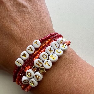 May include: A set of six beaded bracelets with white beads and gold lettering spelling out "I LOVE YOU", "GO", "VA", "HOOK", "TECH", and a heart charm. The bracelets are layered on a person's wrist and are in shades of red, orange, and white.