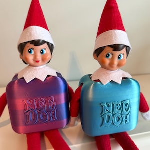 May include: Two elf dolls wearing red and white hats and collars, each encased in a square Nee Doh container. The left doll is in a purple and blue striped container, while the right doll is in a solid light blue container, both with embossed text.