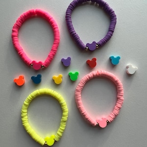 May include: Four colorful bracelets made of stacked plastic beads with a small Mickey Mouse charm on each. The bracelets are in pink, purple, yellow, and light pink. There are also several extra Mickey Mouse charms in various colors.