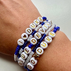 May include: A stack of white beaded bracelets with gold lettering spelling out "I love Duke", "Go Duke", "Blue", and "Devil". The bracelets are on a person's wrist.