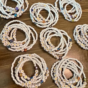 May include: A collection of white beaded bracelets with gold letter beads spelling out words like "MAGIC", "DREAMER", and "DESTINY". Some bracelets feature star-shaped and colorful accent beads. The bracelets are arranged on a wooden surface.