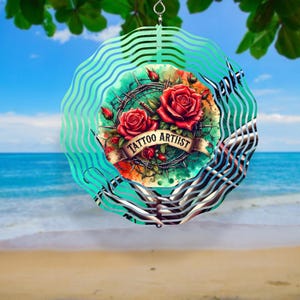 May include: A decorative wind spinner featuring a central design with two red roses, barbed wire, and a banner that reads "TATTOO ARTIST". The spinner has a teal and silver wavy outer design, with a hanging loop at the top.