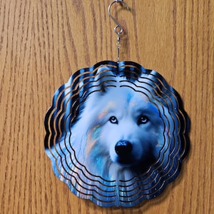 May include: A decorative wind spinner featuring a detailed image of a white wolf with blue eyes. The wolf is surrounded by a layered, wavy, metallic blue and silver frame. The spinner hangs from a metal hook.