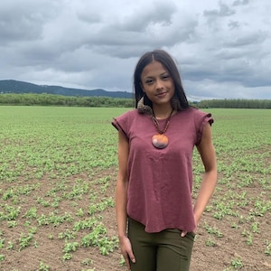 May include: A short-sleeved maroon t-shirt with ruffled sleeve edges, paired with a dark olive green pair of pants and a large, round, multi-colored pendant necklace featuring shades of orange, brown, and black on a dark cord.