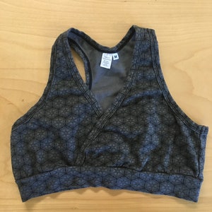 May include: A dark gray racerback sports bra featuring a repeating geometric star pattern in black. The garment has a V-neckline with a wrap-style front, a wide elasticized bottom band, and a white interior label reading Circle Creations with size M.