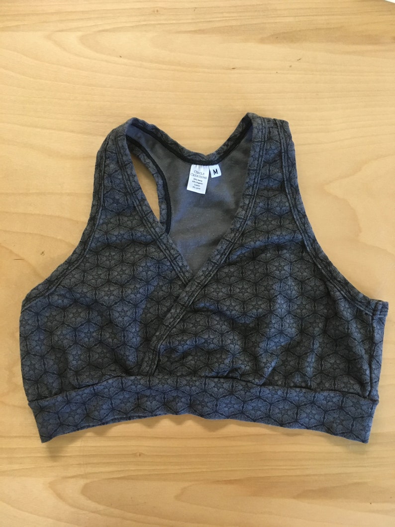 May include: A dark gray racerback sports bra featuring a repeating geometric star pattern in black. The garment has a V-neckline with a wrap-style front, a wide elasticized bottom band, and a white interior label reading Circle Creations with size M.