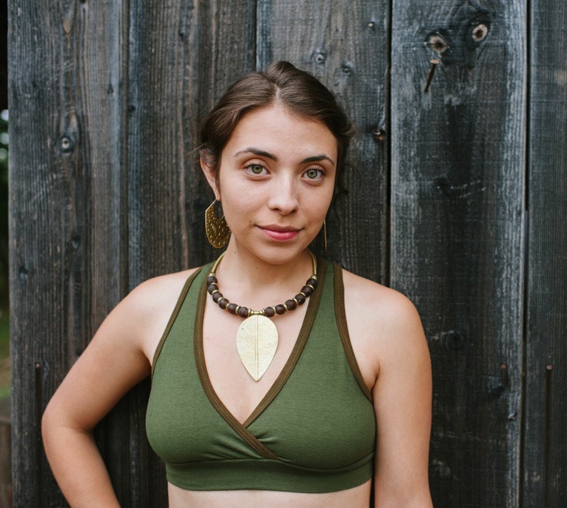 May include: A woman wearing a green, wrap-style sports bra with a brown beaded necklace and a gold leaf pendant. She is standing in front of a wooden wall.