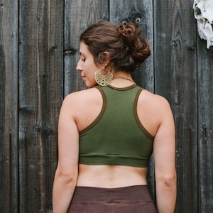 May include: A woman wearing a green racerback tank top with brown trim and a brown skirt. She is standing in front of a wooden wall with a skull hanging on it.