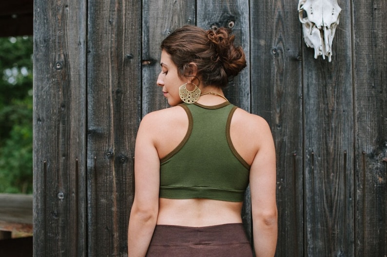 May include: A woman wearing a green racerback tank top with brown trim and a brown skirt. She is standing in front of a wooden wall with a skull hanging on it.