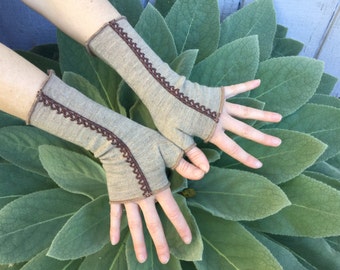 May include: A pair of fingerless gloves in a neutral beige color, trimmed with a dark brown decorative border. The gloves are displayed against a backdrop of large, textured green leaves. The gloves are designed to cover the hand and forearm.