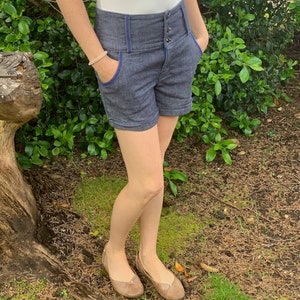May include: High-waisted denim shorts in a dark blue wash with blue trim and pockets. The shorts have a button closure and are worn with a white top and tan flats. The shorts are a stylish and comfortable choice for warm weather.