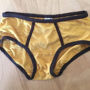 K&ouml;nnte beinhalten: Senfgelbe Baumwollunterw&auml;sche mit braunem Besatz und Spitzendetails. Die Unterw&auml;sche hat einen hohen Bund und eine volle R&uuml;ckenbedeckung.