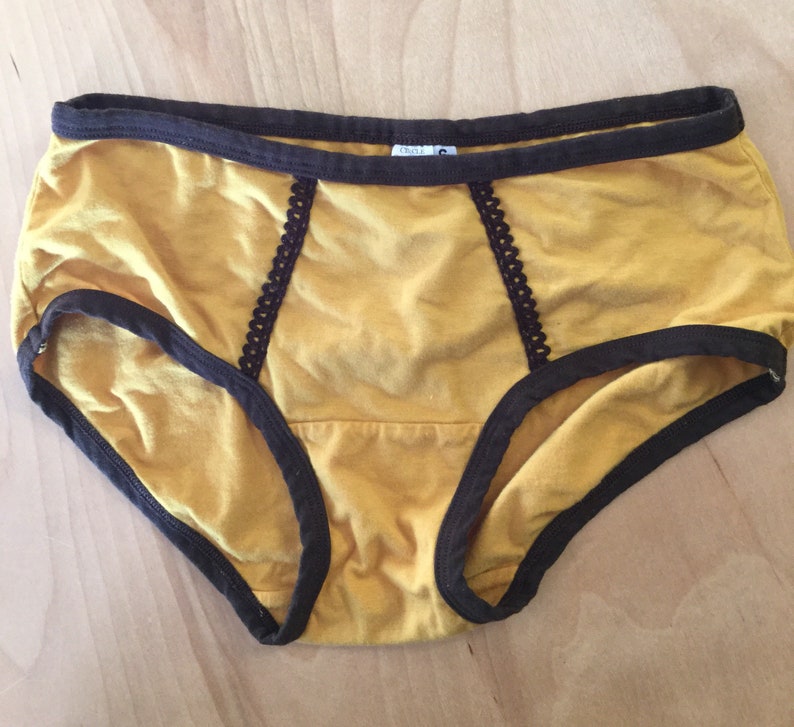 K&ouml;nnte beinhalten: Senfgelbe Baumwollunterw&auml;sche mit braunem Besatz und Spitzendetails. Die Unterw&auml;sche hat einen hohen Bund und eine volle R&uuml;ckenbedeckung.