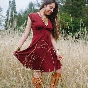 May include: A woman wearing a maroon dress with gold jewelry and brown boots stands in a field of tall grass.