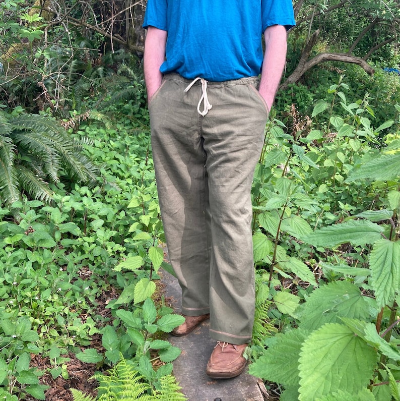 May include: A pair of olive green linen pants with a drawstring waist and a white drawstring. The pants have a relaxed fit and are worn by a person standing in a forest setting.