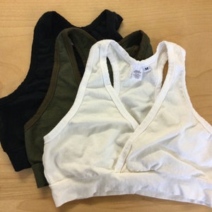May include: A set of three racerback bralettes in white, olive green, and black. Each bralette features a wrap-style front, a wide elasticized bottom band, and a size M tag visible on the white garment. The fabric appears to be a soft cotton blend.