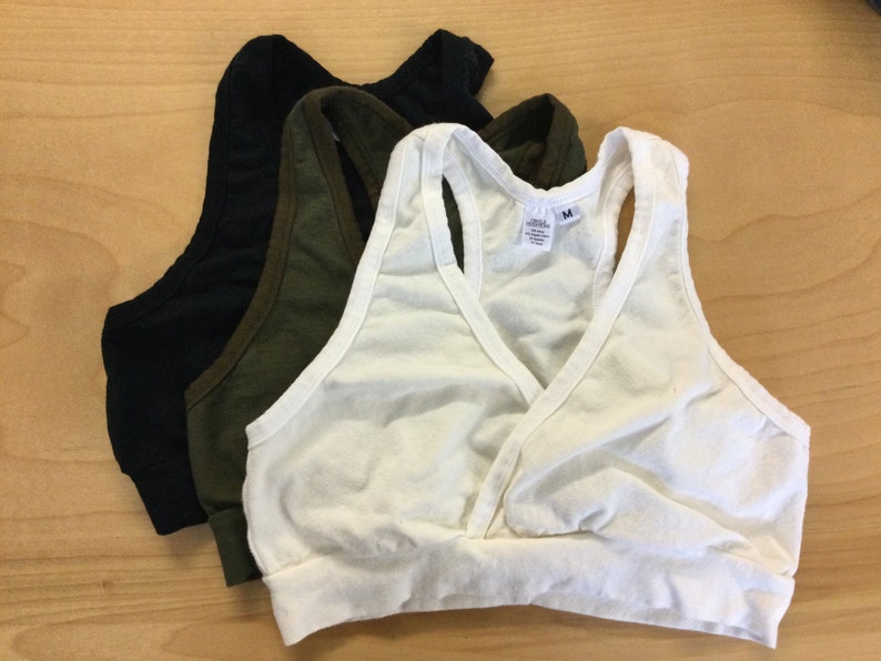 May include: A set of three racerback bralettes in white, olive green, and black. Each bralette features a wrap-style front, a wide elasticized bottom band, and a size M tag visible on the white garment. The fabric appears to be a soft cotton blend.