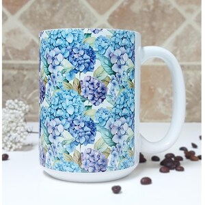 May include: White ceramic mug with a blue and purple floral pattern. The mug has a large handle and is filled with coffee beans.