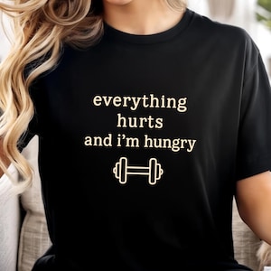 Puede incluir: Camiseta negra con la frase "everything hurts and I'm hungry" en fuente serif, junto con un dibujo de una pesa. La camiseta es de cuello redondo.
