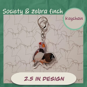 Könnte beinhalten: Ein klarer Acryl-Schlüsselanhänger mit zwei Finken, einer mit orangefarbenen und grauen Markierungen, der andere braun und schwarz. Der Schlüsselanhänger hat einen silbernen Metallring und eine Kette. Der Text auf dem Bild lautet "Society & zebra finch", "Keychain" und "2.5 IN. DESIGN".