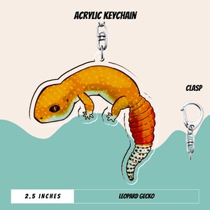 May include: An acrylic keychain featuring a leopard gecko illustration with orange, yellow, and white spotted patterns. The gecko has a thick, banded tail. A silver metal clasp is attached to the top. The text reads ACRYLIC KEYCHAIN, CLASP, 2.5 INCHES, and LEOPARD GECKO.