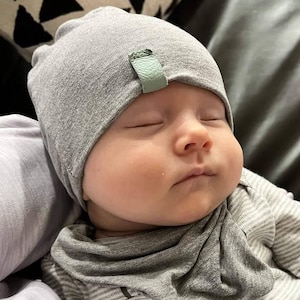 May include: A baby wearing a gray knit hat with a small green tag and a gray striped shirt. The baby is sleeping peacefully.