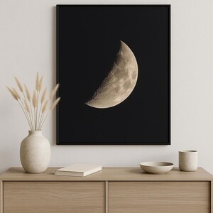 Puede incluir: Una impresión enmarcada que presenta una imagen detallada de una luna creciente sobre un fondo negro. La superficie de la luna está texturizada con cráteres y variaciones de color. La impresión se exhibe sobre un mueble de madera con objetos decorativos.