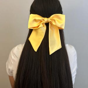 Op de afbeelding: Een grote gele satijnen strik haarclip. De strik is gebonden in een grote, volle knoop en heeft lange, vloeiende staarten.