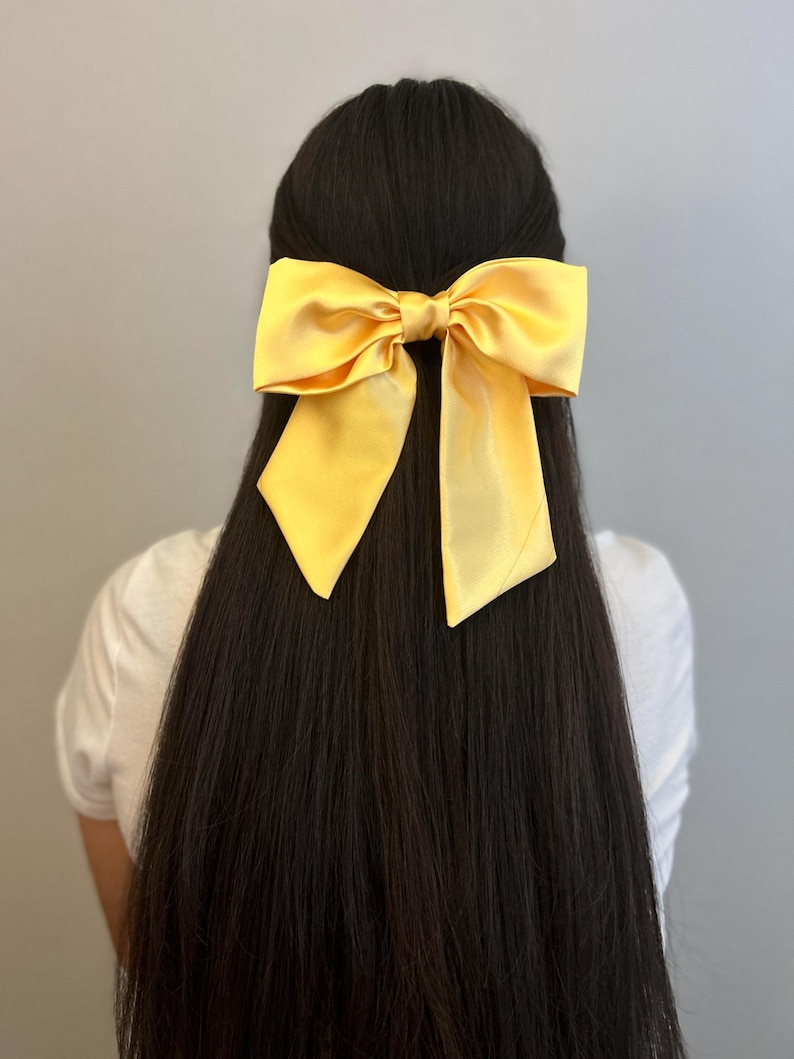 Op de afbeelding: Een grote gele satijnen strik haarclip. De strik is gebonden in een grote, volle knoop en heeft lange, vloeiende staarten.
