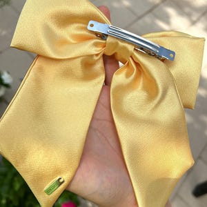 Op de afbeelding: Een grote, gele satijnen strik met een zilveren clip. De strik heeft een klein gouden label met de tekst "Oh Happy Day" erop.