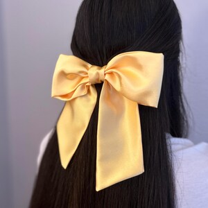 Op de afbeelding: Een grote, gele satijnen strik haarclip. De strik is geknoopt in een grote, symmetrische knoop met lange, vloeiende staarten.