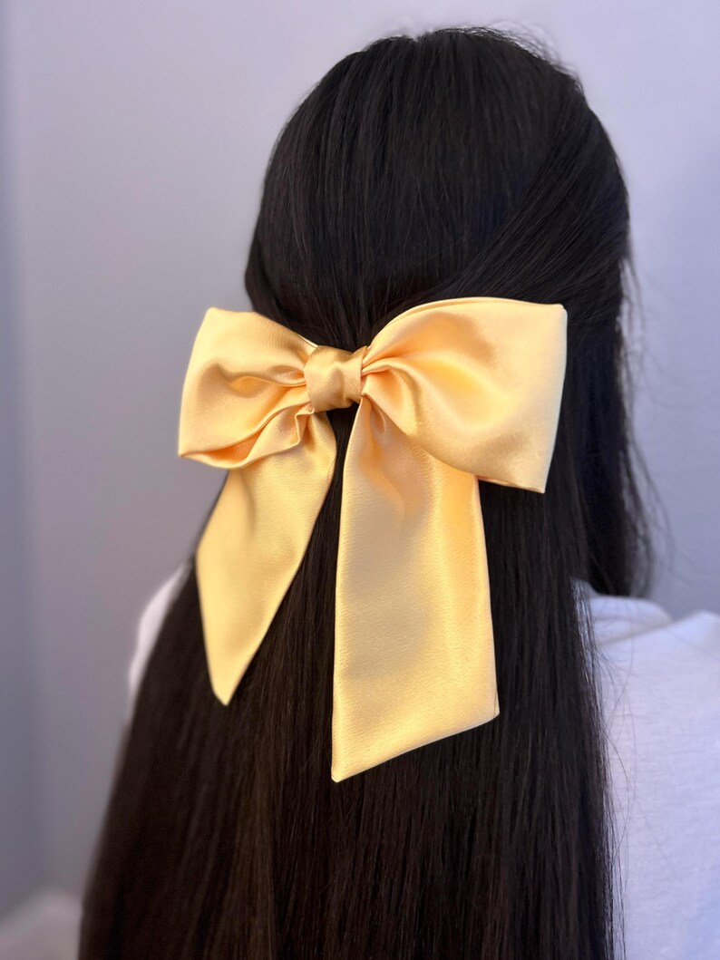 Op de afbeelding: Een grote, gele satijnen strik haarclip. De strik is geknoopt in een grote, symmetrische knoop met lange, vloeiende staarten.
