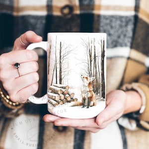 May include: White ceramic mug featuring a watercolor illustration of a fox in a snowy forest scene. The fox is looking up towards the sky. A pile of logs is on the left side of the mug. The mug has a white handle.