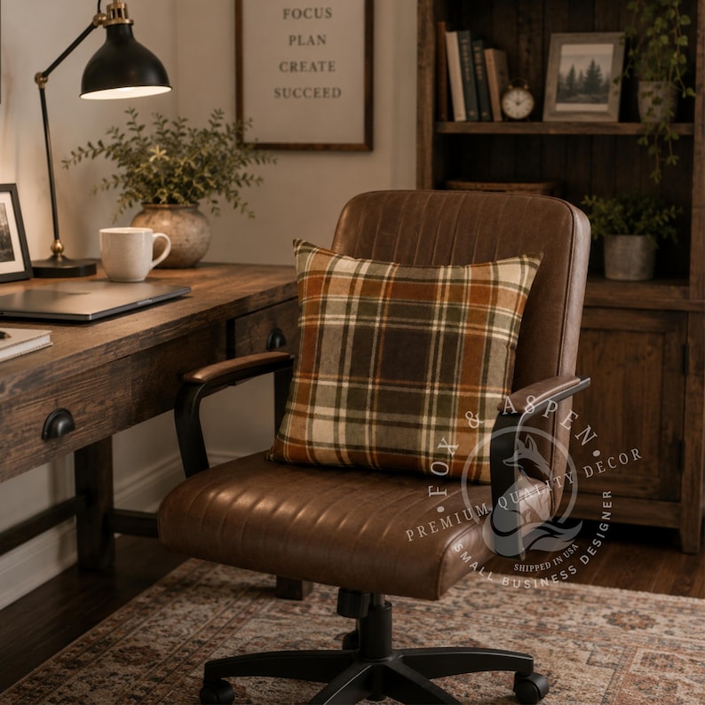 May include: A square throw pillow featuring a plaid pattern in shades of brown, orange, green, and cream. The pillow is placed on a brown leather office chair with black armrests and a five-point base with wheels, set in a home office space.