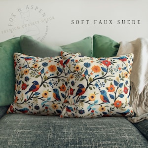 May include: Two decorative throw pillows with a cream-colored background featuring a floral and bird pattern in shades of blue, orange, and yellow. The pillows are labeled "SOFT FAUX SUEDE" and are placed on a gray couch with green pillows.