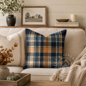 May include: A square throw pillow featuring a classic plaid pattern in shades of navy blue, tan, and brown. The fabric has a soft texture and is displayed prominently in the center, showcasing a grid of intersecting horizontal and vertical lines.
