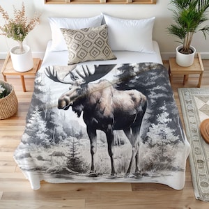 May include: A white and grey throw blanket featuring a black and white illustration of a moose standing in a snowy forest. The moose has large antlers and is looking to the left.