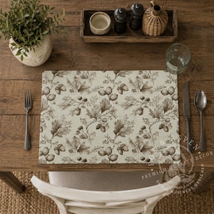 May include: A rectangular placemat featuring a repeating botanical pattern of brown oak leaves and acorns on a light cream background. The placemat is positioned on a wooden table with a fork, a knife, a spoon, and a clear glass nearby.