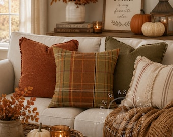 May include: Four square throw pillows arranged on a light-colored sofa. One is textured rust orange, one features a green and orange plaid pattern, one is solid olive green, and one is cream with vertical brown stripes and fringed edges.