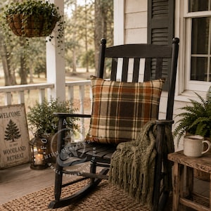 May include: A black wooden rocking chair featuring a square throw pillow with an orange, brown, and cream plaid pattern. A textured olive green knit throw blanket with fringe is draped over the seat, resting against the back of the chair.