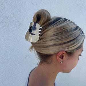 May include: A beige plastic hair claw with a guitar design. The claw is holding a woman's blonde hair in a bun.