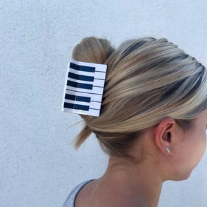 Puede incluir: Una pinza para el pelo blanca con un diseño de teclas de piano negras. La pinza está en el pelo de una persona con cabello rubio.