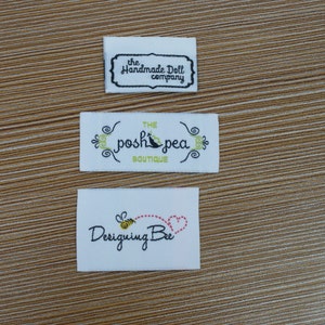 Puede incluir: Tres etiquetas de tela blanca con texto negro y de color. La etiqueta superior dice "the Handmade Doll company" en una fuente decorativa. La etiqueta del medio dice "THE posh pea BOUTIQUE" con detalles florales. La etiqueta inferior dice "Designing Bee" con una abeja y un coraz&oacute;n.