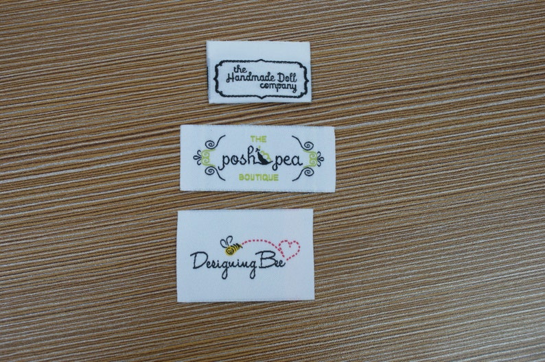 Puede incluir: Tres etiquetas de tela blanca con texto negro y de color. La etiqueta superior dice "the Handmade Doll company" en una fuente decorativa. La etiqueta del medio dice "THE posh pea BOUTIQUE" con detalles florales. La etiqueta inferior dice "Designing Bee" con una abeja y un coraz&oacute;n.