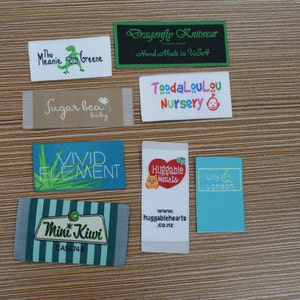 Puede incluir: Una colecci&oacute;n de etiquetas de tela con varios nombres de marcas y logotipos. Las etiquetas incluyen "The Melanie Greene" con un dinosaurio, "Dragonfly Knitwear", "Sugar bea baby", "Vivid Element", "Mini Kiwi", "ToodaLouLou Nursery", "Huggable Hearts" y "Lily & London".