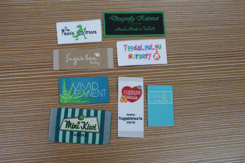 Puede incluir: Una colecci&oacute;n de etiquetas de tela con varios nombres de marcas y logotipos. Las etiquetas incluyen "The Melanie Greene" con un dinosaurio, "Dragonfly Knitwear", "Sugar bea baby", "Vivid Element", "Mini Kiwi", "ToodaLouLou Nursery", "Huggable Hearts" y "Lily & London".